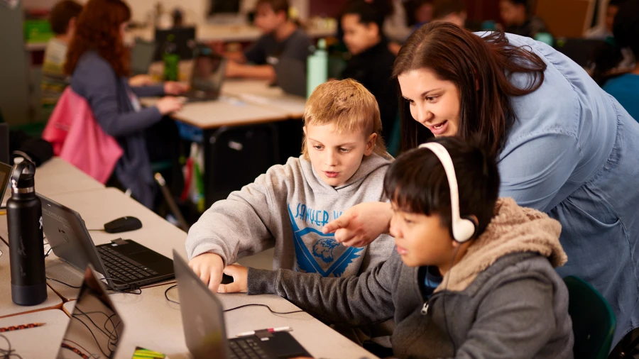 Um professor se inclina sobre dois alunos em uma mesa, ajudando-os com o Minecraft Education.